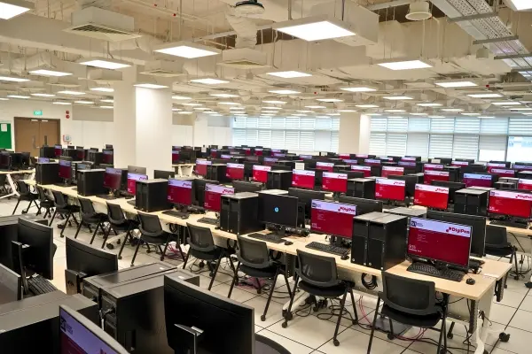 Students sit at rows upon rows of computer monitors and keyboards in a large fluorescent-light-lit computer lab.