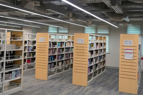 A library with shelves of books and desks. A window looks out to a tree outside.